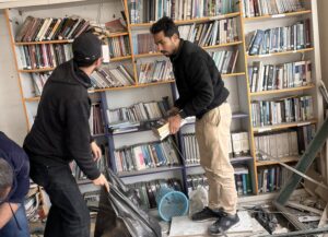 Dua pemuda sedang mengevakuasi buku-buku dari rak ke dalam kantong plastik hitam besar di dalam ruangan perpustakaan yang hancur dipenuhi puing-puing bangunan dan atap yang runtuh.
